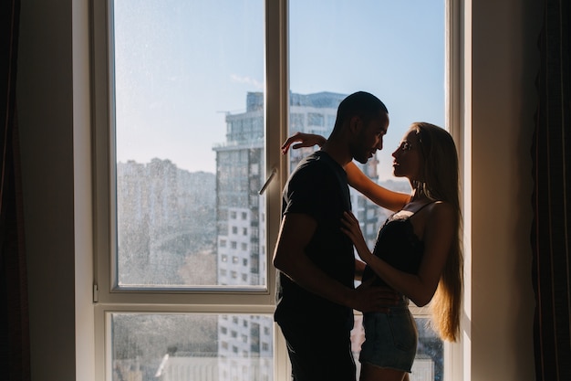 真の素敵な感情を表現し、近代的なアパートで晴れた朝の窓の近くを抱く若いカップルの情熱。関係、愛、レジャー、自宅で、長いブロンドの髪を持つ魅力的な女性