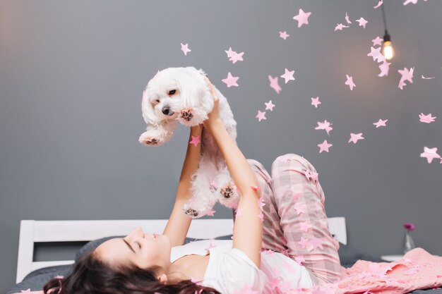 Expressing true positive emotions of young joyful woman in pajamas with brunette curly hair having fun with little dog in falling pink tinsels on bed