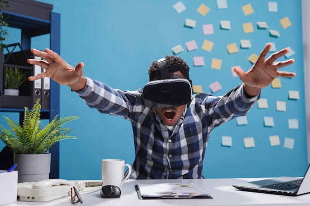 Expressive amazed african american company office employee enjoying virtual reality gaming technology. Adult businessman wearing electronic entertainment device while sitting at desk in workspace.