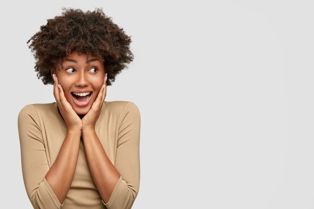 Expressive attractive African American female exclaims from happiness, keeps hands on cheeks, looks joyfully aside as notices best friend returning from abroad, stands against white wall, blank space