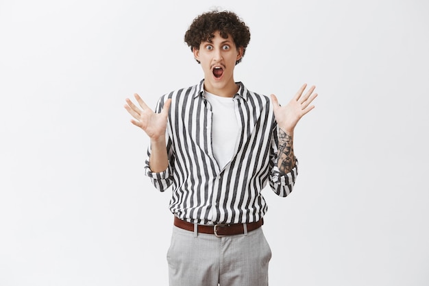 Expressive excited and amazed attractive stylish urban hipster male with curly hair and moustache in trendy striped blouse and pants gesturing with palms and opened mouth explaining stunning story