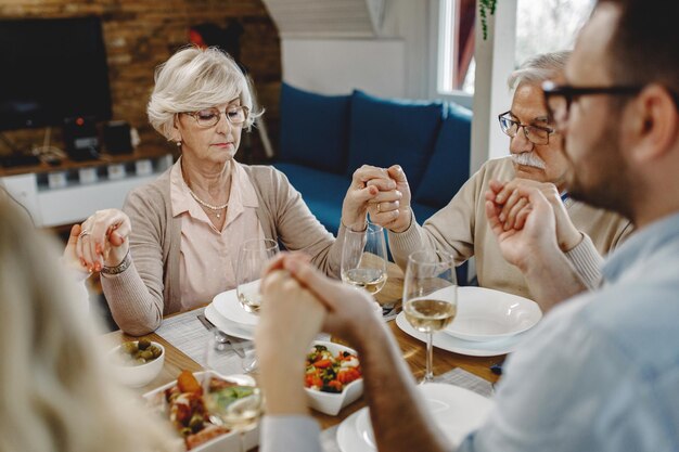 식탁에서 손을 잡고 은혜를 말하는 대가족은 노인 여성에게 초점을 맞춥니다.