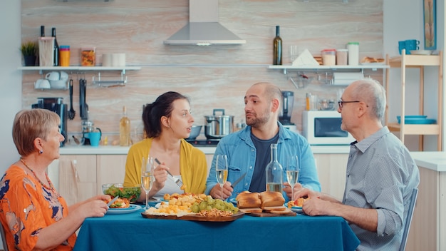 Extended family talking having relaxing time. Multi generation enjoying time at home, in the kitchen sitting by the table, eating dinner together and drinking