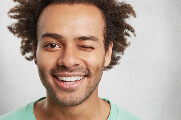 Extreme close up of mixed race handsome man has trendy hairdo, smiles pleasantly and blinks eye