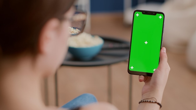 Extreme closeup of young woman holding vertical smartphone with green screen watching online video content or webinar in living room. Girl using touchscreen mobile phone with chroma key in video call.