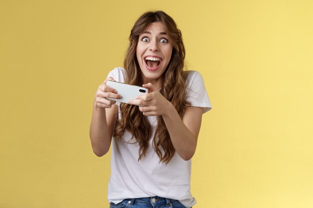 Extremely happy thrilled playful girl gamer playing awesome great new smartphone game hold mobile phone horizontal cheering look camera astonished impressed beating record yellow background