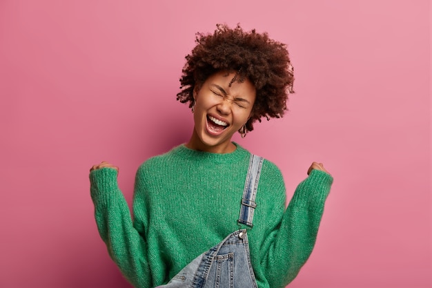 Free photo extremely joyful dark skinned woman raises clenched fists, exclaims from happiness, feels lucky to win something, celebrates amazing victory, closes eyes and smiles broadly, isolated on pink wall