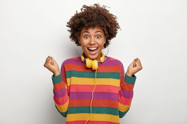 Free photo extremly happy african american lady celebrates scored goal, makes victory gesture, raises clenched fists, has curly hairstyle, wears striped colorful jumper, uses headphones connected to some device