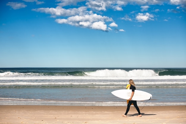 Eye-level shot of a blonde female with a surfboard walking barefoot along the seashore