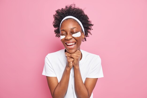 Eye skin care. Cheerful young Afro American woman keeps hands under chin smiles broadly applies collagen patches for removing fine lines 