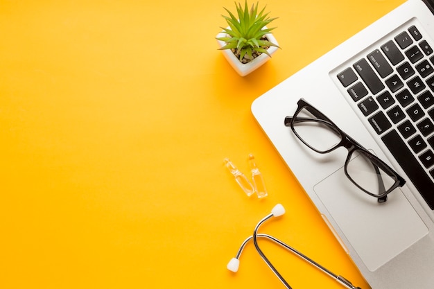 Free photo eyeglass over laptop with ampoule; stethoscope with succulent plant against yellow backdrop