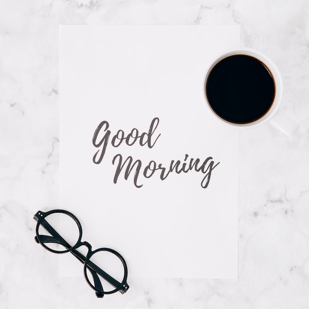 Eyeglasses and coffee cup over the good morning text on paper over white marble textured backdrop