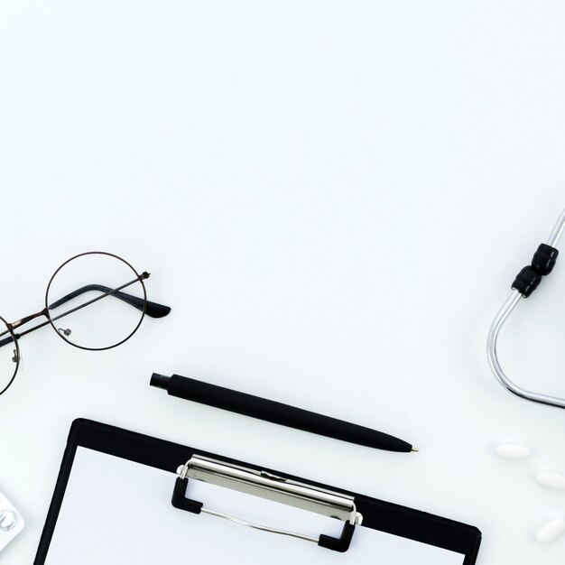 Eyeglasses; pen; clipboard; pills and stethoscope on white backdrop