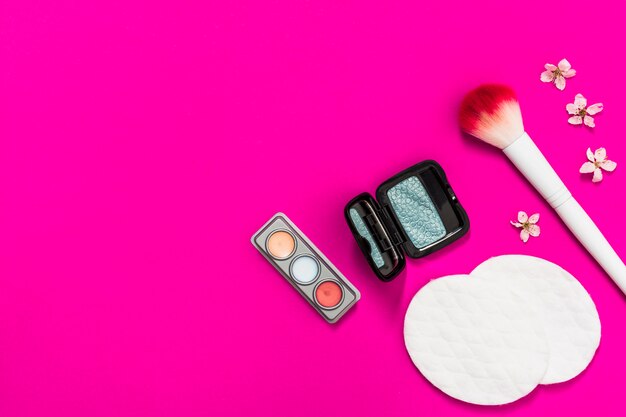 Eyeshadow palette; makeup brush; cotton pads and flower on pink background