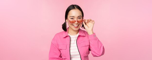 Eyewear advertisement Stylish modern asian girl touches sunglasses wears pink poses against studio background Copy space