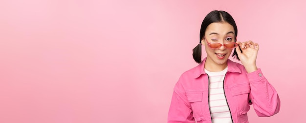 Eyewear advertisement Stylish modern asian girl touches sunglasses wears pink poses against studio background Copy space