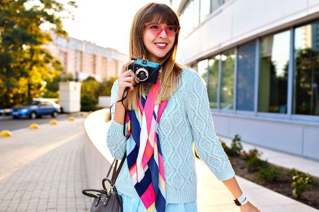 Fabulous young stylish woman posing on the street, romantic elegant outfit, sweater scarf and sunglasses, holding vintage camera end enjoying time.