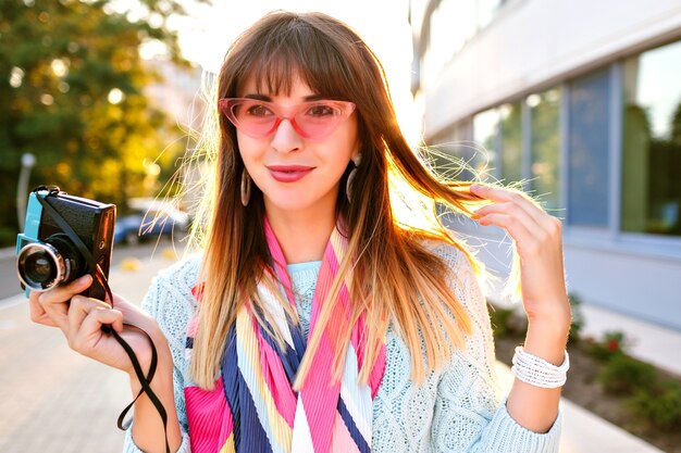 Fabulous young stylish woman posing on the street, romantic elegant outfit,  sweater scarf and sunglasses, holding vintage camera end enjoying time.