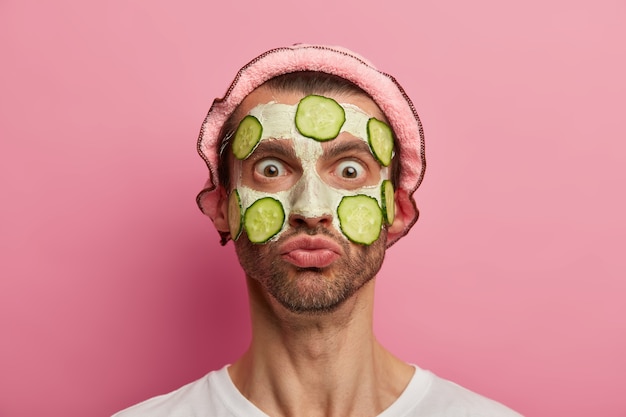Face care concept. Shocked emotive man stares with bugged eyes at his reflection, applies white clay mask with slices of cucumber
