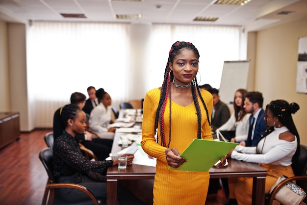 Лицо красивой африканской деловой женщины в желтом платье, держащей буфер обмена на фоне встречи многорасовой команды деловых людей, сидящей за офисным столом