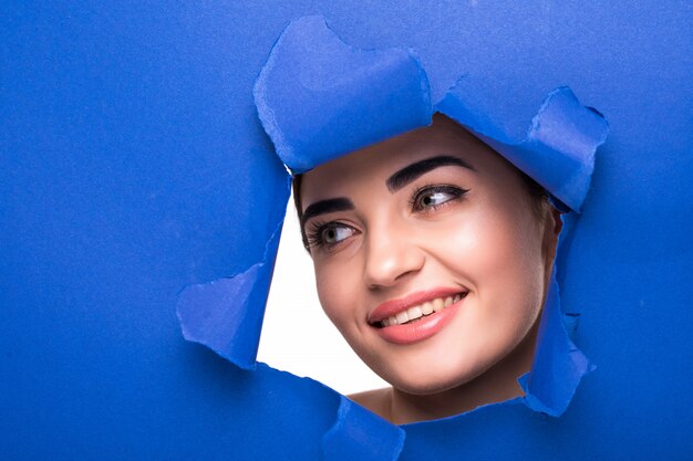 The face of a young beautiful woman with a bright make-up and puffy blue lips peers into a hole in blue paper.