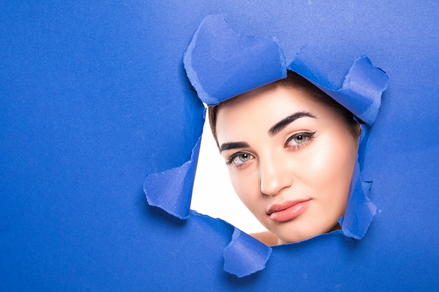 The face of a young beautiful woman with a bright make-up and puffy blue lips peers into a hole in blue paper.