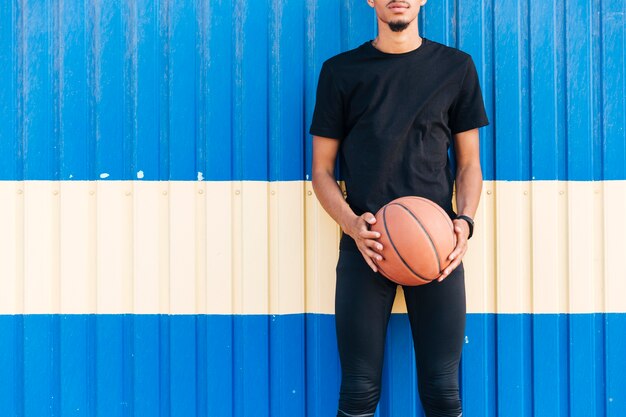 Безликий атлетический мужчина стоя против стены держа баскетбол