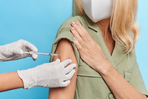 Бесплатное фото Безликая блондинка в защитной маске, платье получает прививку в плечо для профилактики болезней, позирует на фоне синей стены