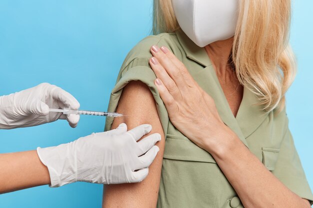 Faceless blonde woman wears protective face mask dress gets inoculaton in shoulder for disease prevention poses against blue wall