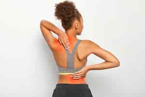 Free photo faceless curly woman suffers from spine pain, wears sport bra, shows location of inflammation, isolated on white background.