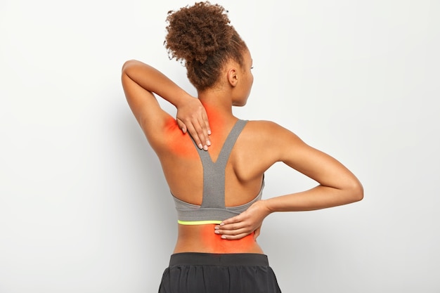 Faceless curly woman suffers from spine pain, wears sport bra, shows location of inflammation, isolated on white background.