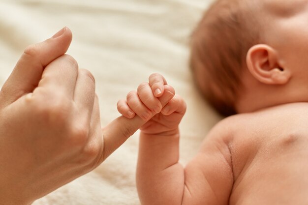 Faceless mother with naked baby, infant holding mommy's finger, mum spending time with her tiny child on light background.