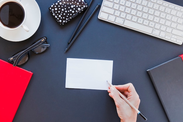 Faceless person writing on note near stationery and keyboard on desk with coffee cup