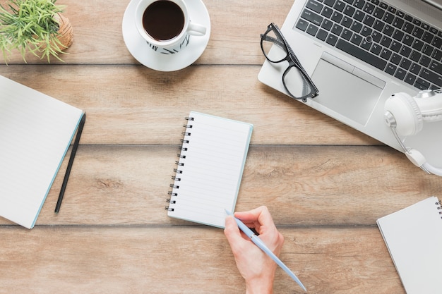 Faceless person writing in notebook near stationery and laptop on table