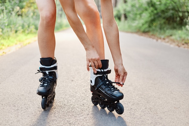 Faceless portrait of woman lace up roller skates while rollerblading outdoor in summer park on asphalt road, unknown female person rollerskating alone.