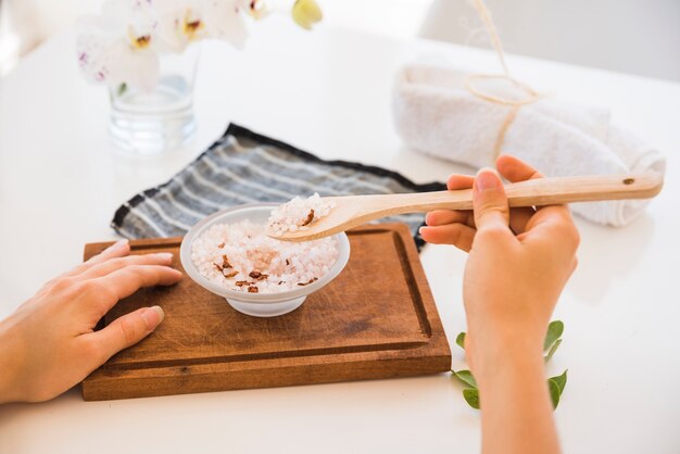 Faceless woman with spoon and pink salt in bowl on cutting board