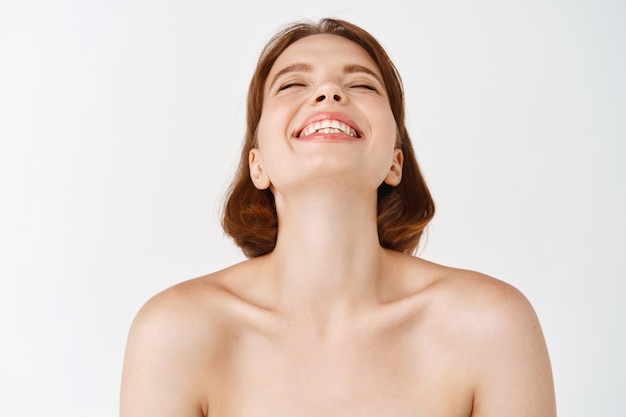 Foto gratuita bellezza del viso. ragazza naturale con pelle sana e pulita, spalle nude, ridente e sorridente spensierata. donna libera che si gode una sensazione fresca e pulita sul viso, muro bianco