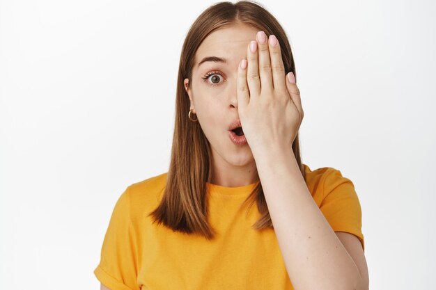 Facial expressions and people. Girl looks surprised with one eye, cover half of face with hand, gasp and say wow impressed, checking out big sale, white background
