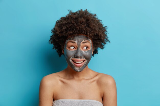 Facial treatment concept. Positive curly haired woman applies clay mask on face to rejuvenate skin undergoes beauty procedures after taking shower poses wrapped in towel isolated over blue wall