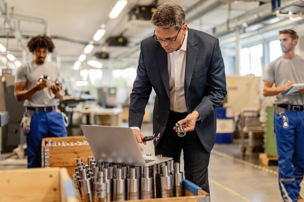 Factory manager reading instructions on a computer while checking measurements of steel bolt in industrial building Manual workers are in the background