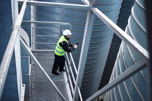 Lavoratore di silos di fabbrica in piedi sulla piattaforma metallica tra i serbatoi di stoccaggio industriali e guardando la tavoletta sulla produzione alimentare