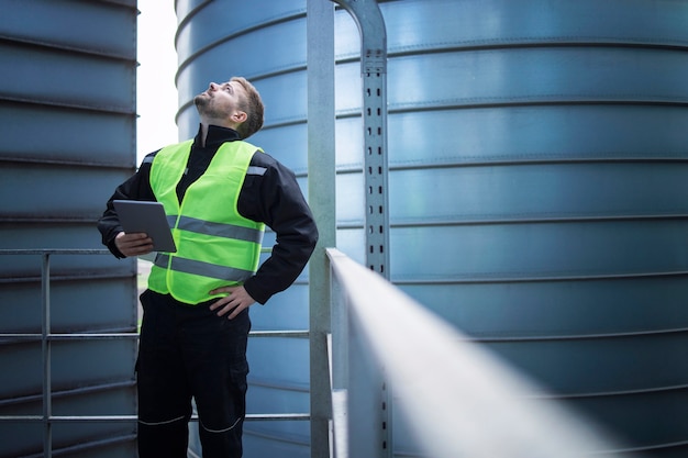 Factory worker standing on metal platform between industrial storage tanks and looking up for visual inspection of silos for food production