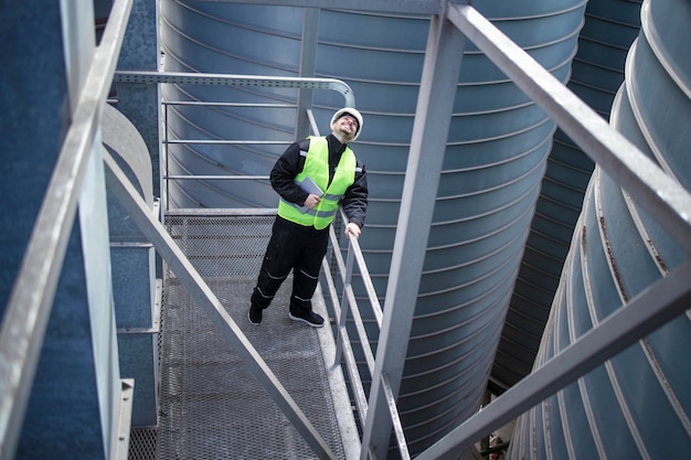 Factory worker standing on metal platform between industrial storage tanks and looking up for visual inspection of silos food production