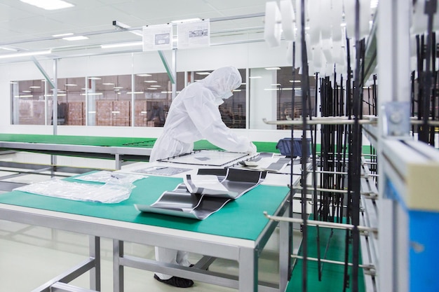 Фабричный рабочий в белом лабораторном костюме и маске для лица собирает части телевизора на столе