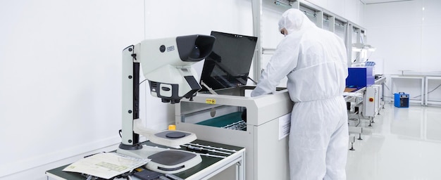 A factory worker in a white lab suits and face masks working with some modern equipment in a clean white room an electron microscope is in the foreground