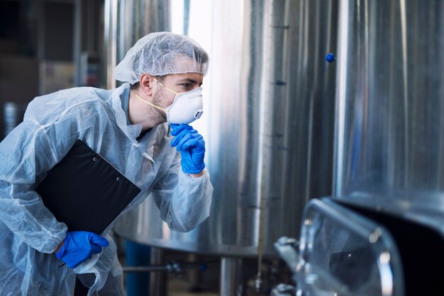 Factory worker in white protective uniform with hairnet and mask looking at parameters of an industrial machine