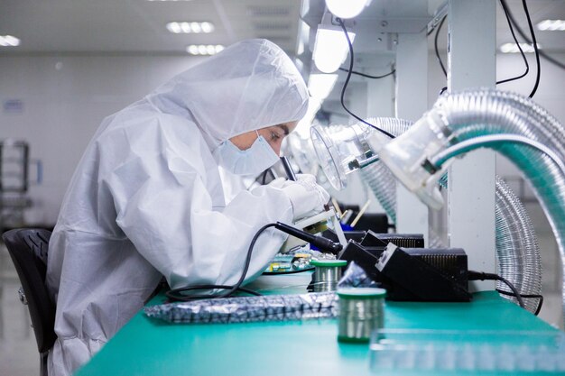 Factory workers in white lab suits and face masks sitting on the chairs are producing tv sets with soldering irons under bright lamps