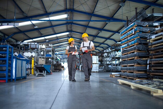 Factory workers in work wear and yellow helmets walking through industrial production hall and discussing about delivery deadline