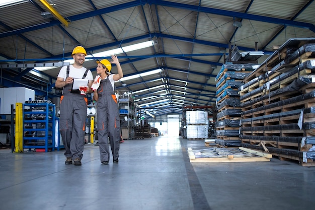 Factory workers in work wear and yellow helmets walking through industrial production hall and discussing about organization