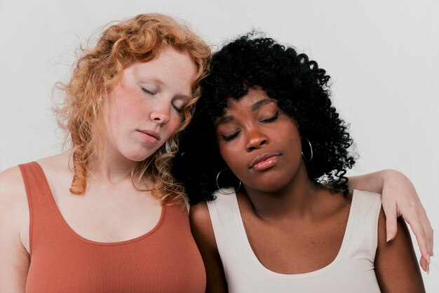 Fair and dark skin women leaning to each other sleeping against grey background